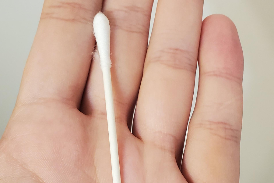 A close-up of a hand holding a single cotton swab with a wooden stick and white cotton tip, set against a neutral background.