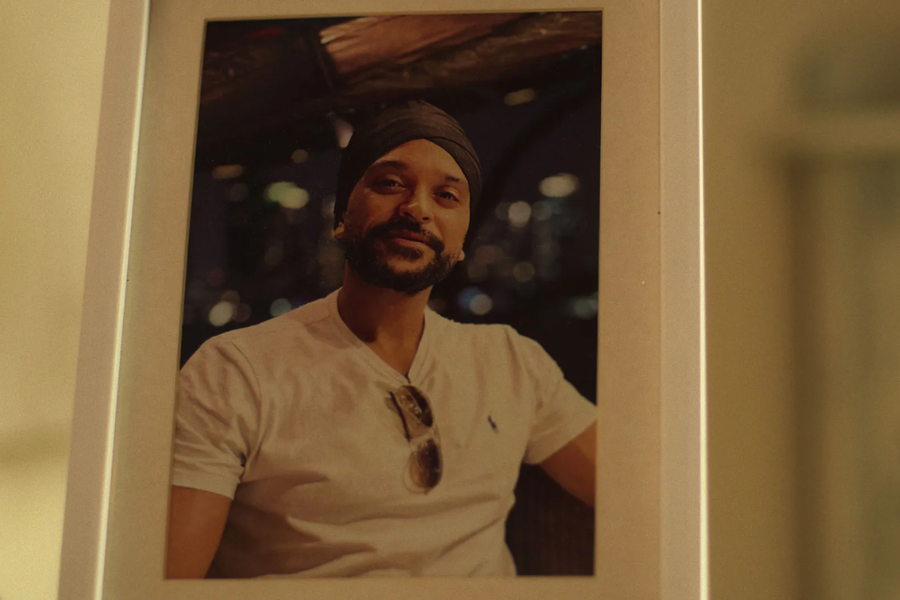 A framed photograph of a man wearing a white t-shirt, sunglasses clipped to his collar, and a dark turban, smiling slightly while seated against a blurry nighttime cityscape backdrop.