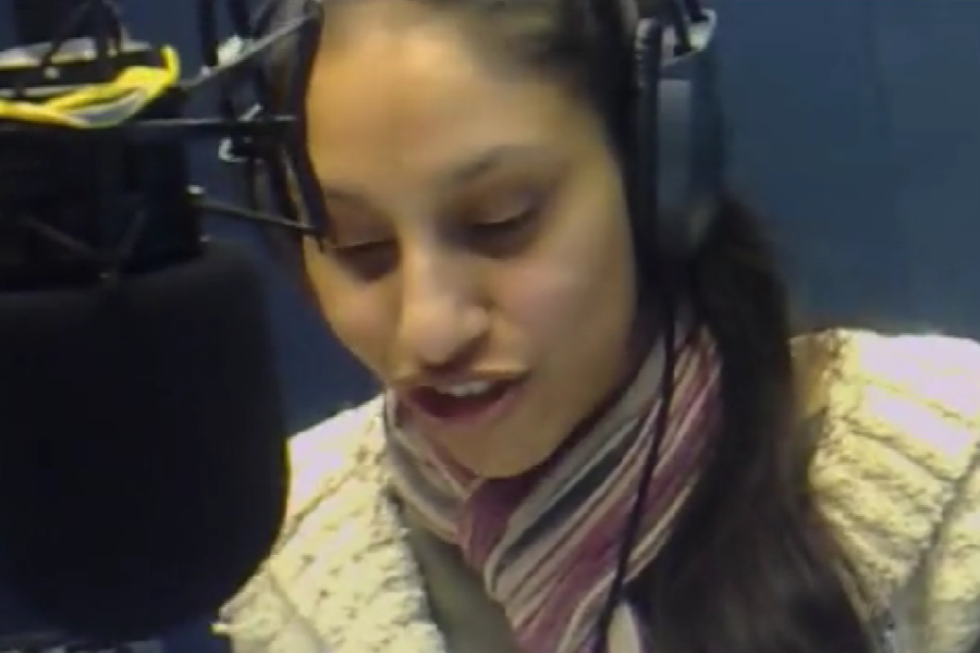 A woman wearing large headphones and a striped scarf speaks into a professional microphone in a radio studio. She appears focused, possibly reading or announcing something live on air.