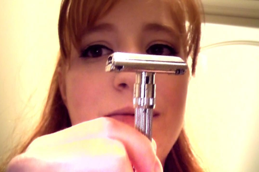 A woman with red hair holds up a silver safety razor in front of her face, focusing on the razor while standing in a bathroom.