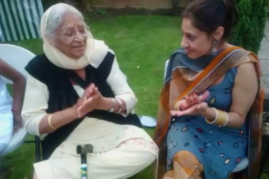 An elderly woman and a younger woman sit outdoors on white chairs, clapping and smiling at each other. The older woman wears a cream outfit with a black vest and headscarf, while the younger woman is dressed in a blue and orange salwar kameez.