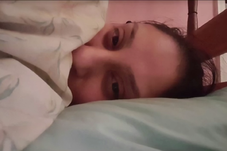 A woman lies in bed with her face partially covered by a floral blanket, peeking out with tired eyes and resting her head on a light blue pillow.