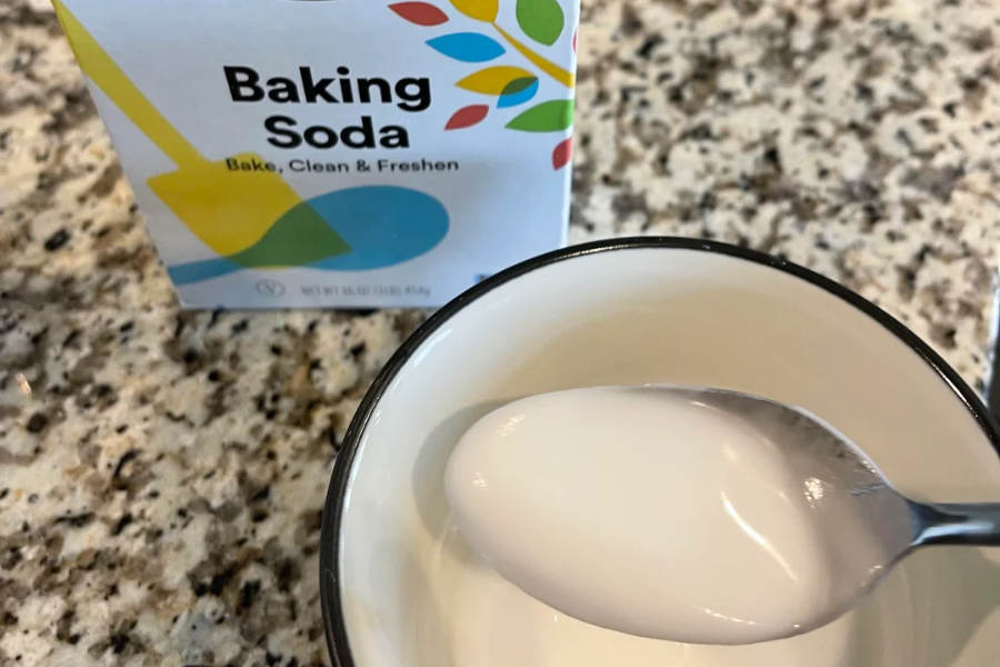 A box of baking soda sits on a granite countertop next to a bowl containing a thick white paste, with a spoon lifting some of the mixture, likely made from baking soda and water.
