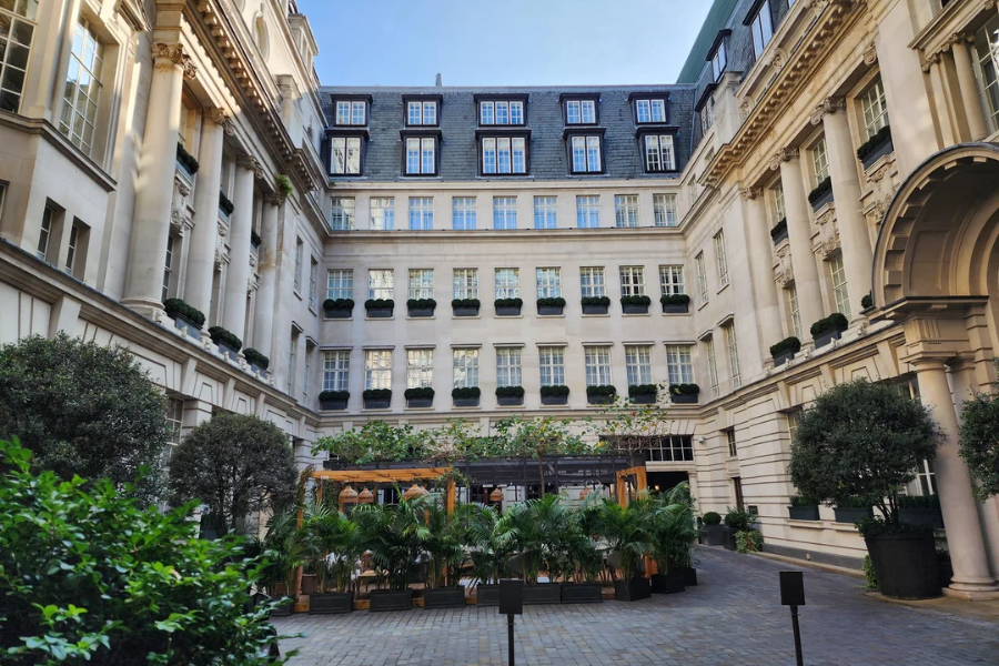 A grand courtyard surrounded by elegant multi-story buildings with classical architectural details and rows of uniform windows. In the center, lush greenery and a wooden pergola structure hint at an upscale outdoor dining or lounge area.