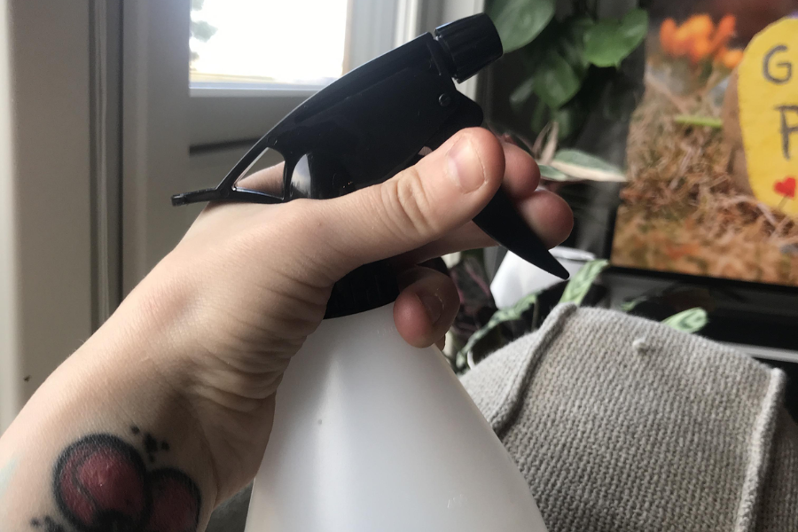A hand with a heart tattoo holding a white spray bottle with a black nozzle, with indoor plants and a cozy living room scene visible in the background.