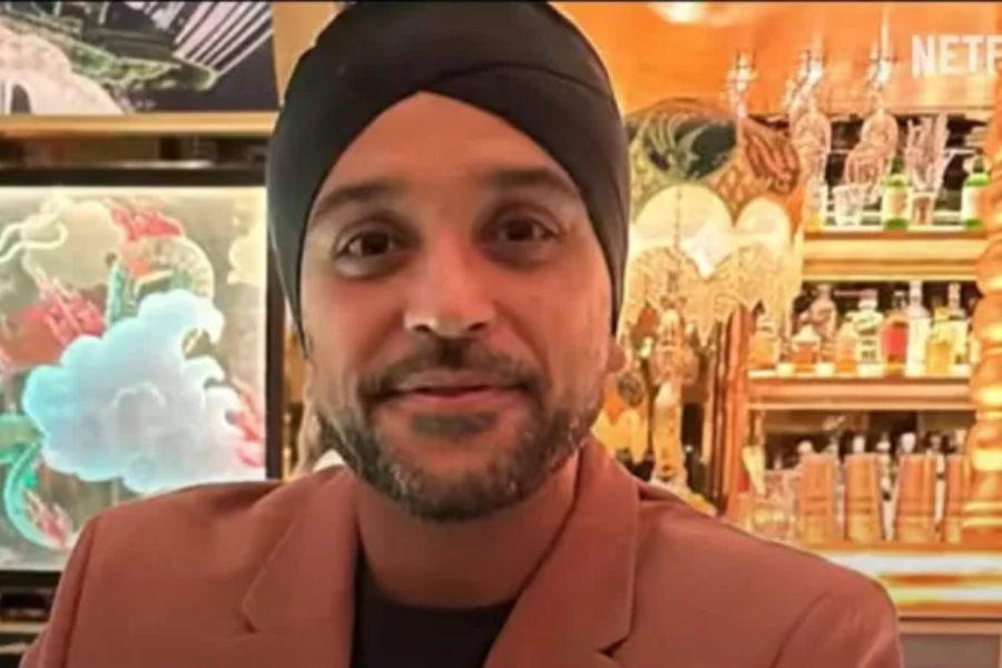 A man wearing a black turban and a brown blazer stands in front of a well-lit bar filled with bottles and ornate décor. The word "Netflix" is partially visible in the top right corner, suggesting a scene from a show or documentary.