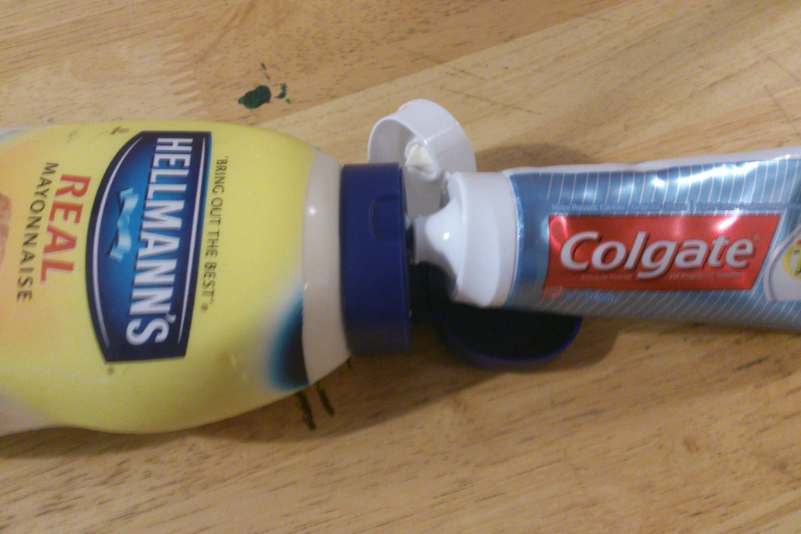 A humorous setup showing a Colgate toothpaste tube squeezed into the opening of a Hellmann’s Real Mayonnaise bottle, both lying on a wooden surface with their caps open.