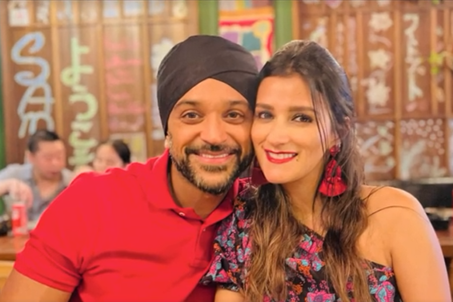 A smiling couple poses closely together in a lively restaurant setting. The man wears a black turban and red polo shirt, while the woman wears a colorful floral dress with red earrings and bold red lipstick. The background is filled with people and wall decorations with Asian characters.