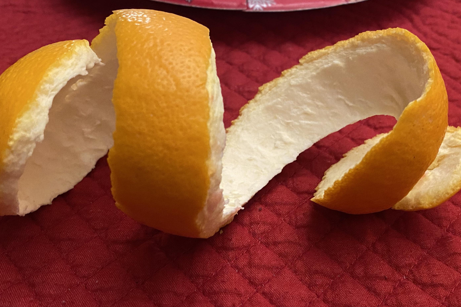 A long, continuous strip of orange peel sits curled on a red quilted surface, with the white pith visible on the inner side of the peel.