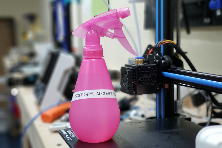 A bright pink spray bottle labeled "ISOPROPYL ALCOHOL 99%" sits on the bed of a 3D printer in a workspace with various electronic tools and components in the background.