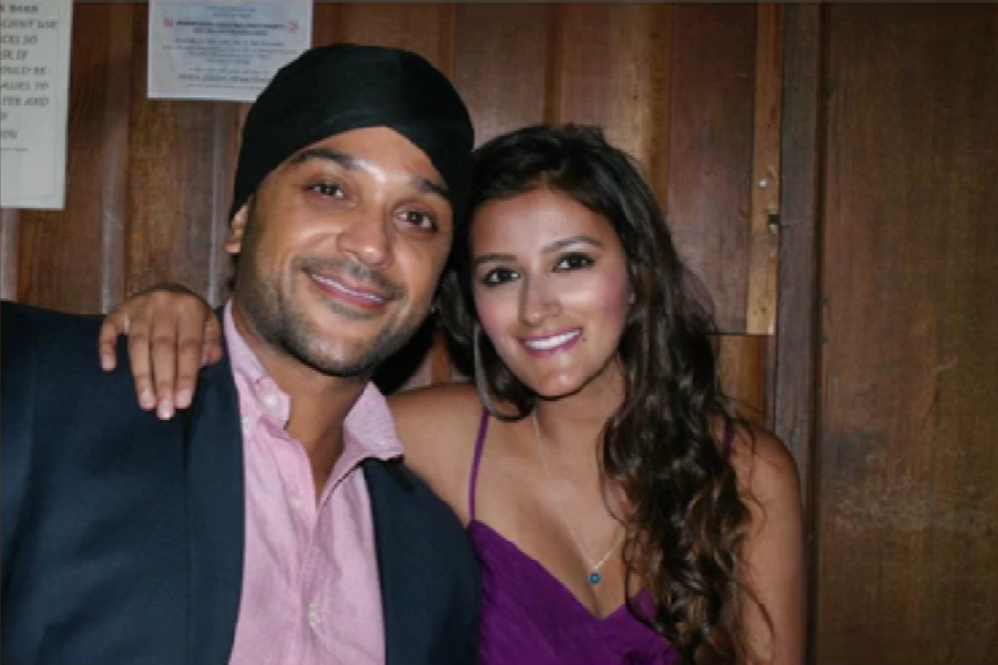 A smiling couple poses closely together in front of a wooden wall. The man wears a black turban, navy blazer, and light pink shirt, while the woman has long wavy hair and is dressed in a purple spaghetti strap dress with a pendant necklace.