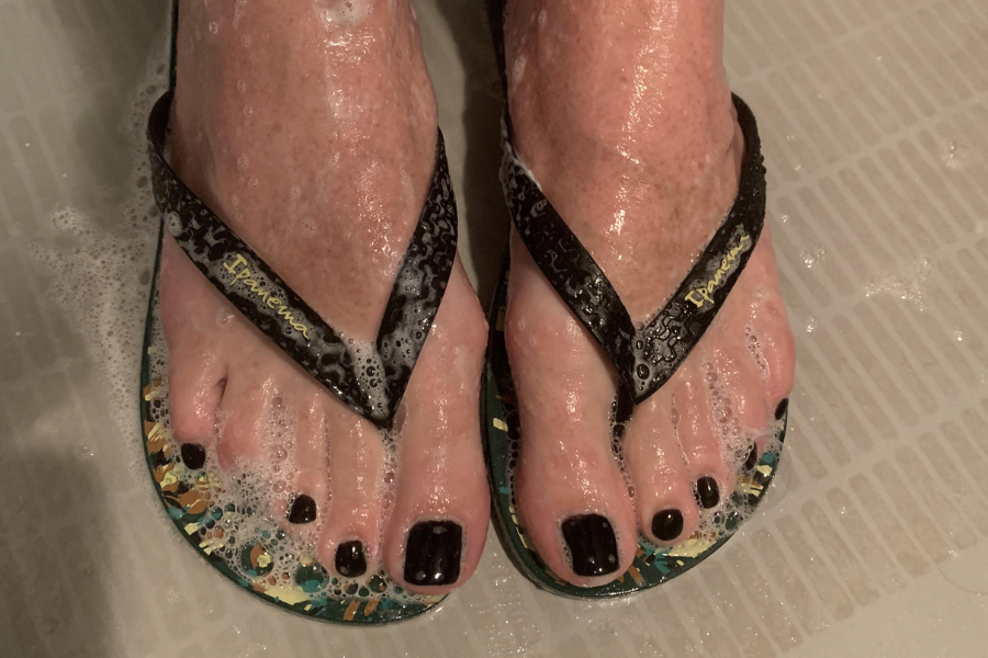 A pair of wet feet wearing flip-flops with black straps and colorful soles, standing on a tiled shower floor. The toes have glossy black nail polish, and soap suds are visible around the feet.