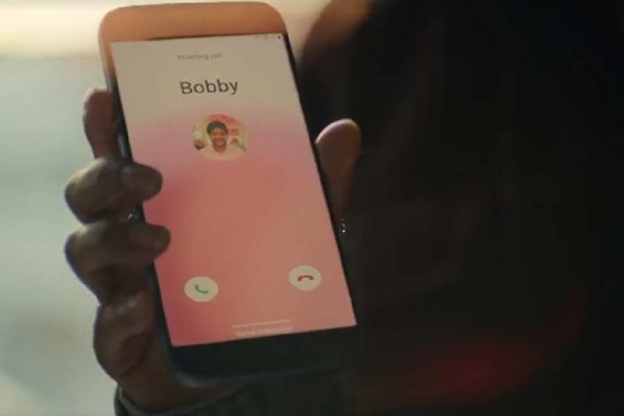  A hand holds a smartphone displaying an incoming call from “Bobby” with a pink gradient screen. The caller's photo shows a smiling man, and green and red buttons offer options to accept or decline the call.