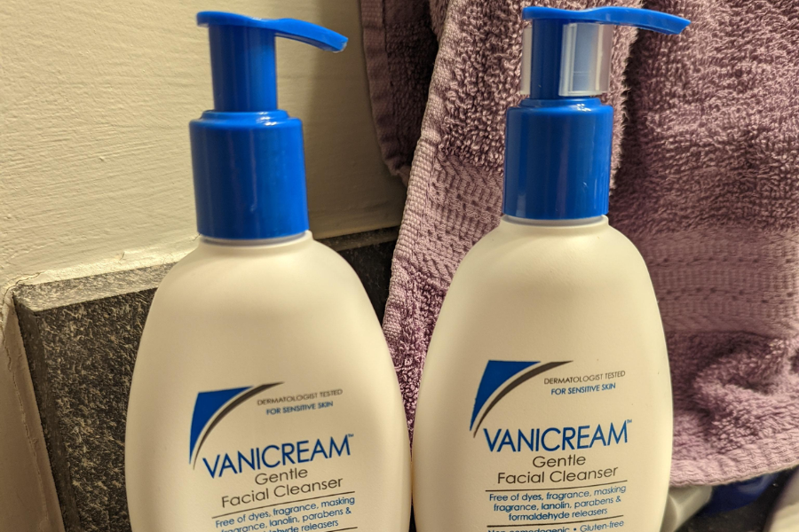 Two bottles of Vanicream Gentle Facial Cleanser with blue pump tops are placed on a bathroom counter in front of a purple towel. The labels highlight that the product is free of dyes, fragrance, lanolin, parabens, and formaldehyde.