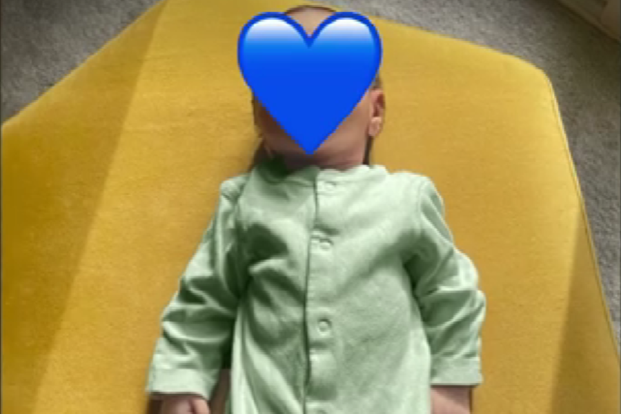 A baby dressed in a light green onesie lies on a bright yellow cushion. The baby’s face is covered with a large blue heart emoji for privacy.