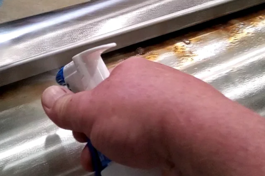 A hand holding a spray bottle aimed at a rusted area on a corrugated metal surface, likely applying a cleaning or rust-removal solution.