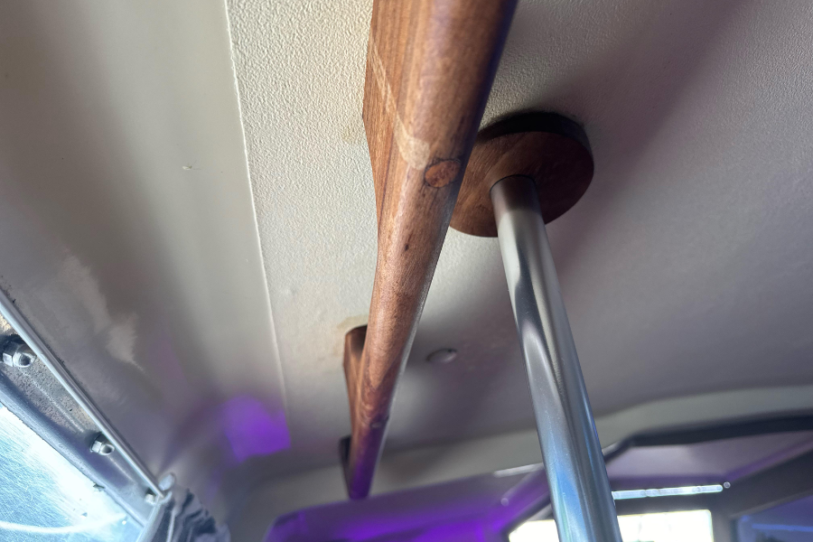 Close-up view of a wooden handle or handrail mounted to the ceiling of a vehicle interior, supported by a round wooden bracket and a metal pole, with windows and purple ambient lighting visible in the background.