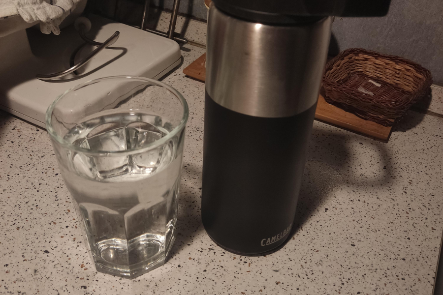 A glass filled with water sits next to a black and stainless steel CamelBak thermos on a speckled kitchen countertop, with various kitchen items in the background.