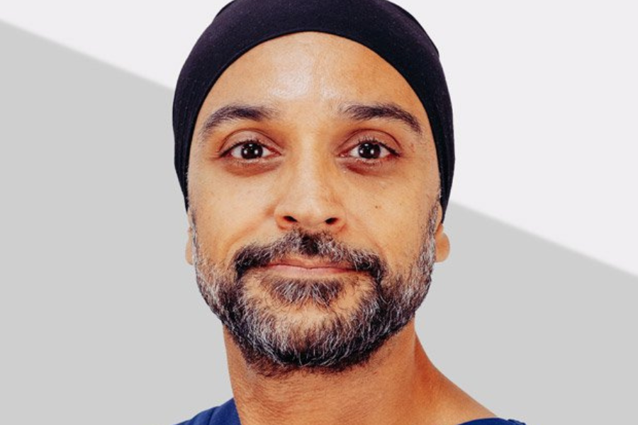 A portrait of a man with a neatly groomed beard and mustache, wearing a black turban and a navy shirt. The background is a simple, two-tone grey and white design, giving the image a professional look.