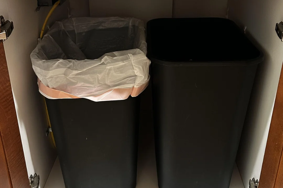 Two black rectangular trash bins are placed side by side inside a cabinet under a sink; the left bin has a white trash bag lining, while the right bin is unlined.