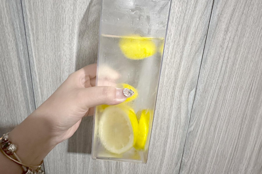 A hand with a glittery ring and charm bracelet holds a tall, rectangular water bottle filled with ice water and several lemon slices, against a light wood-grain background.