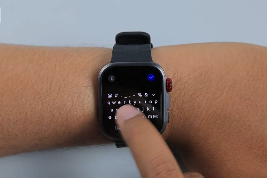 A person uses their index finger to type on a QWERTY keyboard displayed on a smartwatch screen. The smartwatch has a black strap and a red crown, and the keyboard includes letters, symbols, and a blue check mark icon in the corner.