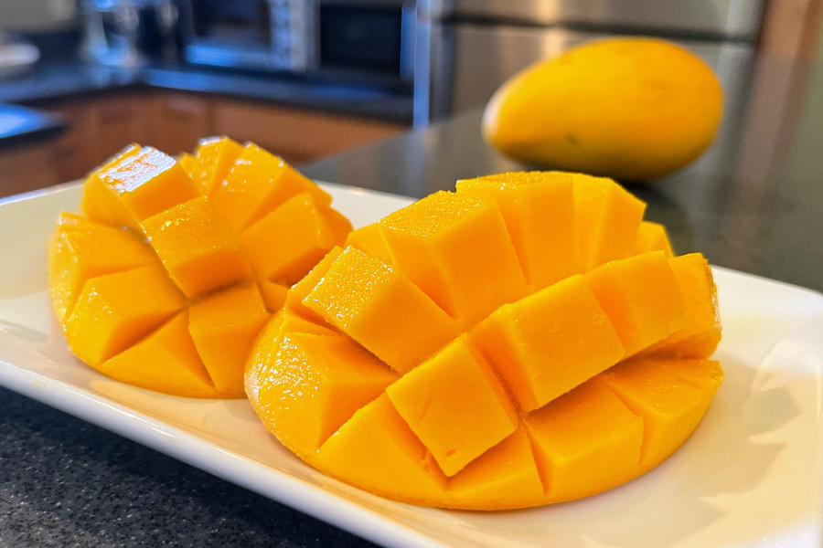 Two mango halves cut into a grid pattern and turned inside out, revealing vibrant yellow-orange cubes of fruit on a white plate. A whole mango sits blurred in the background on a kitchen counter.