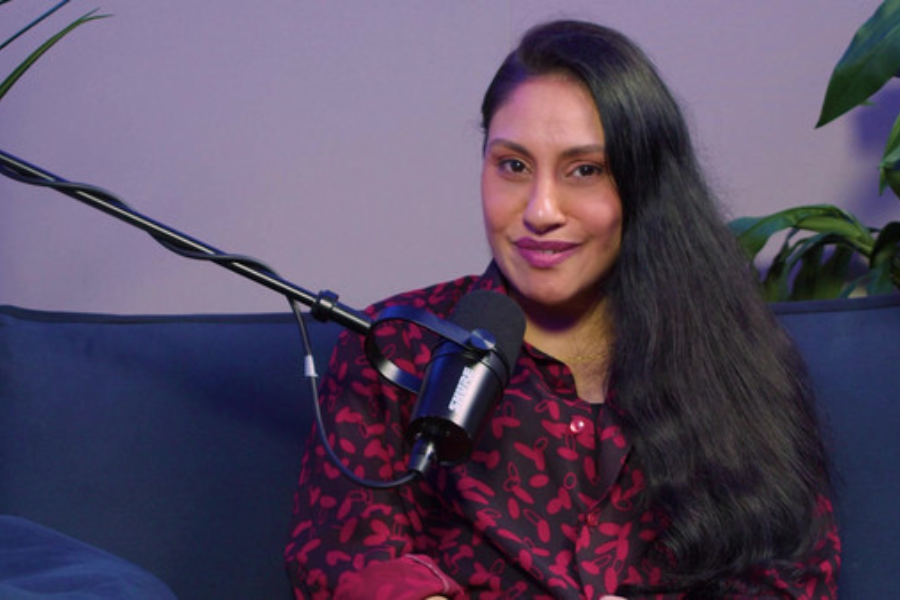 A woman with long dark hair sits on a blue couch in front of a microphone, wearing a red and black patterned blouse, smiling slightly during a podcast or interview recording session with plants in the background.