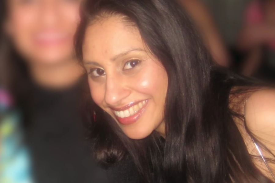 A woman with long, straight dark hair smiles brightly at the camera in a softly lit setting, with a blurred crowd in the background suggesting a social gathering.