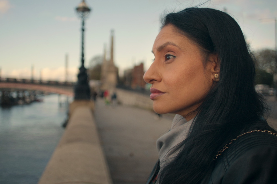 A woman with long dark hair stands by a riverside walkway, gazing thoughtfully into the distance with a bridge and lampposts lining the background under a clear sky.