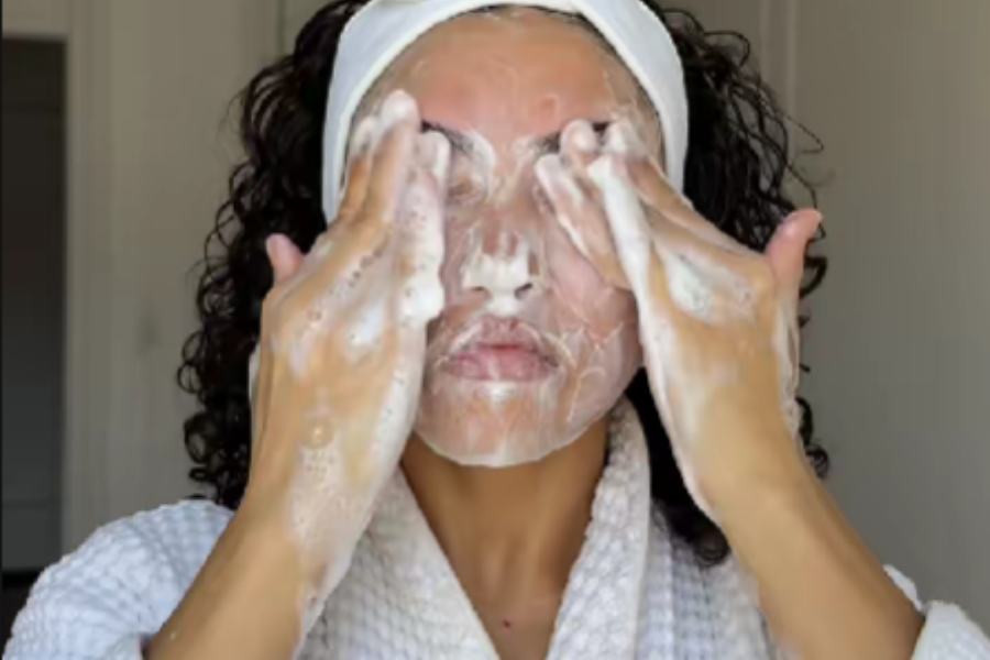 A person wearing a white headband and bathrobe is cleansing their face with a foamy facial cleanser, using both hands to gently massage the product around the eye and cheek area.