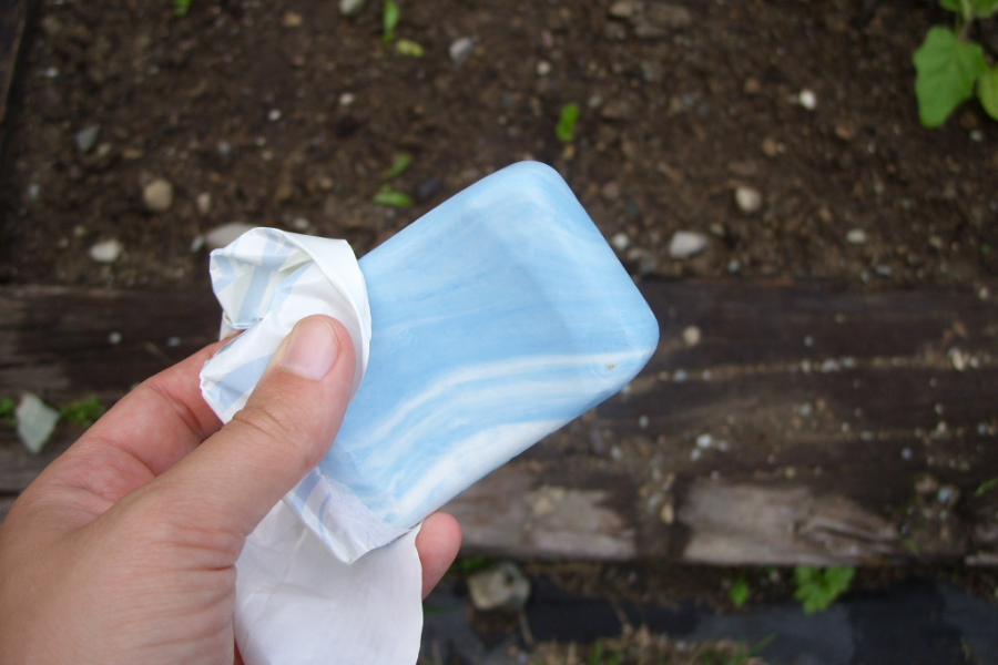 A hand holding an unwrapped bar of blue and white marbled soap over a garden bed with soil and small green sprouts.