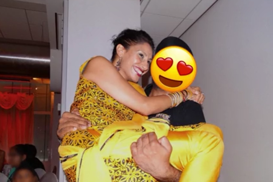 A woman in a yellow traditional outfit is being joyfully lifted in the arms of a person whose face is covered with a heart-eyes emoji. Both appear to be dressed for a festive occasion, smiling and surrounded by soft lighting and guests in the background.