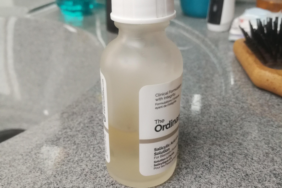 A partially used bottle of The Ordinary Salicylic Acid 2% Solution sits on a bathroom counter, with surrounding toiletries and a hairbrush in the background.