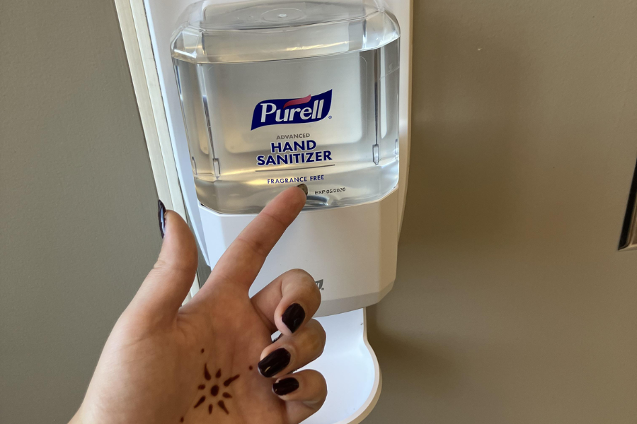 A hand with dark nail polish and a sun-shaped henna design is pressing a wall-mounted Purell Advanced Hand Sanitizer dispenser labeled “Fragrance Free.”