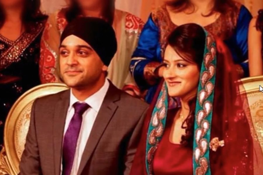 A man in a dark suit and turban sits beside a woman in traditional South Asian attire, wearing a deep red outfit with an embroidered dupatta, surrounded by guests in vibrant clothing, suggesting a wedding or engagement ceremony.